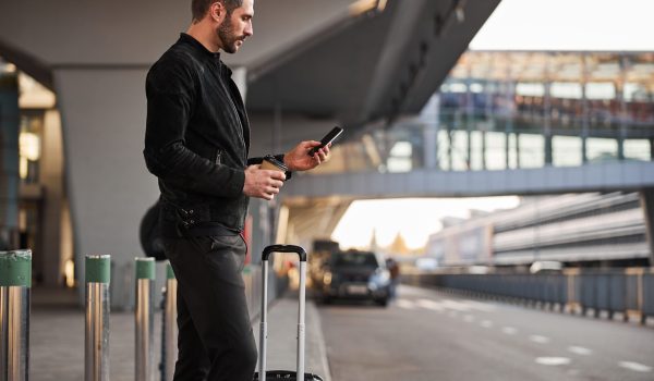 Calm handsome man with a wheeled suitcase drinking coffee outside the airport and looking at the cellphone screen